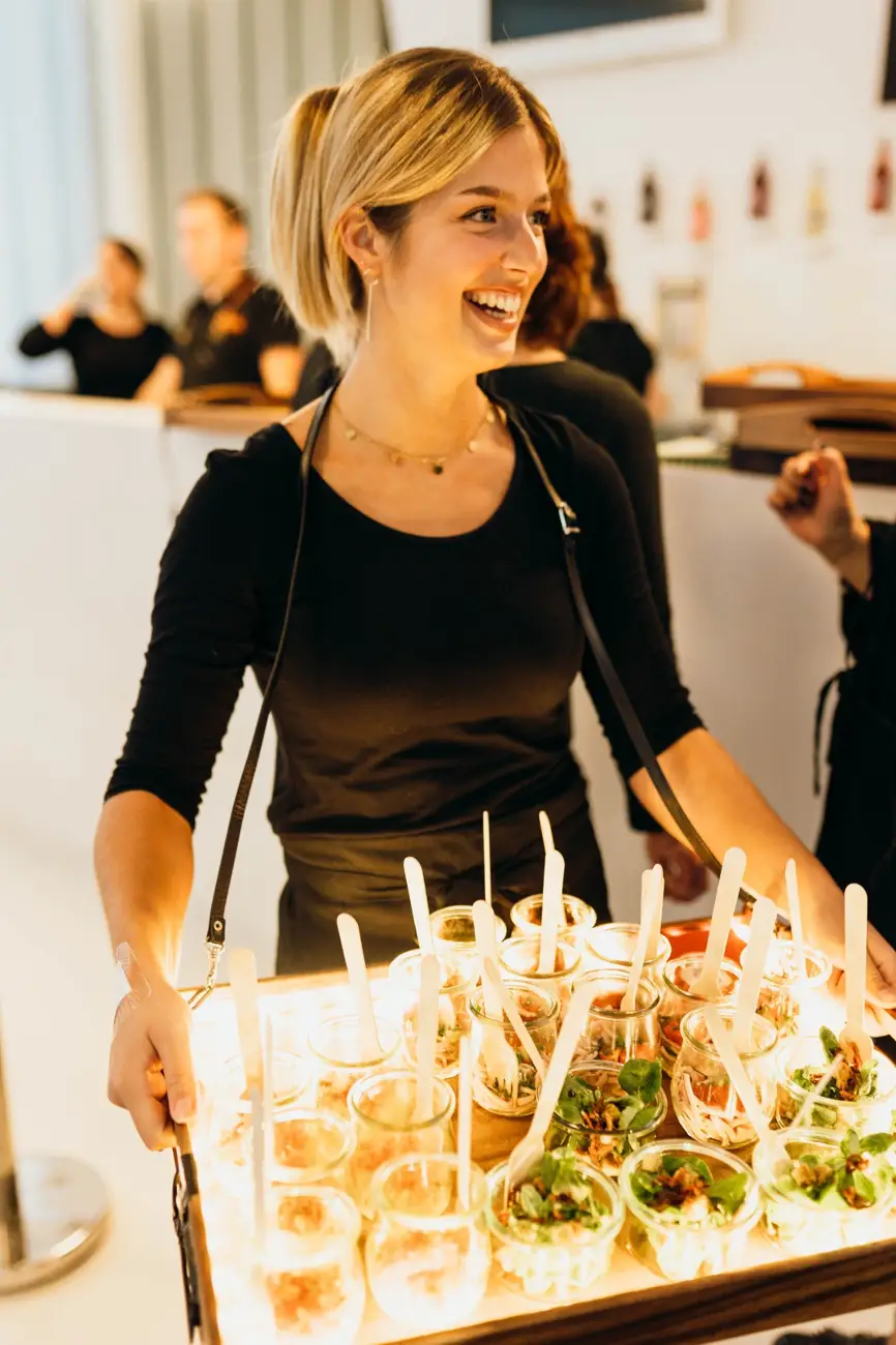Servicekraft trägt ein beleuchtetes Tablett mit Salat- und Fingerfood-Gläschen durch die Gäste